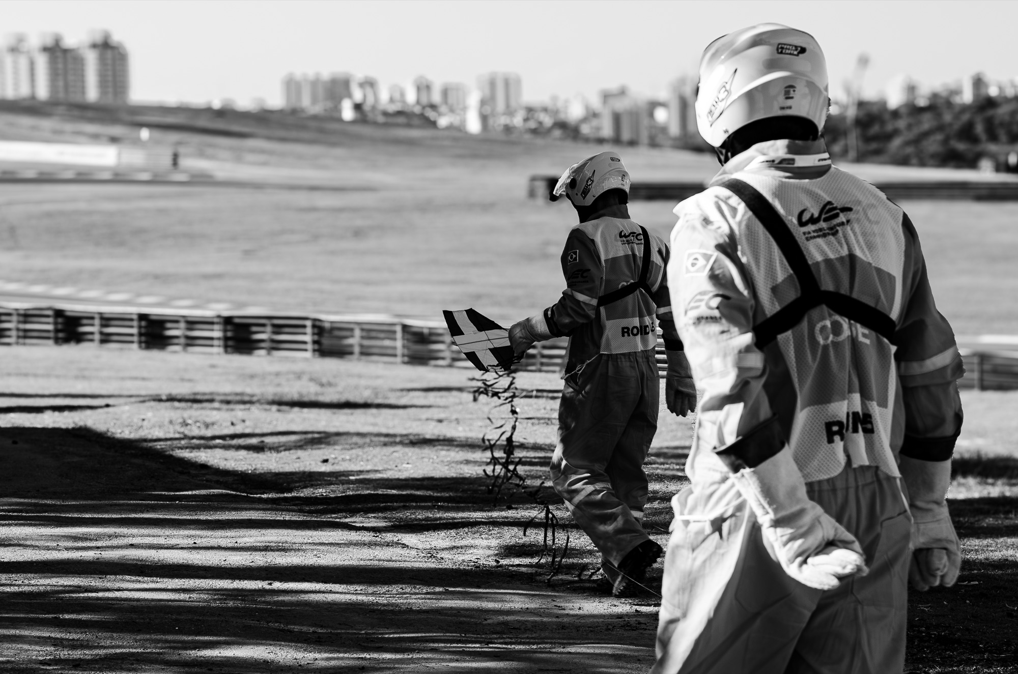 Dois fiscais do WEC recolhendo destroços na área de escape, com a cidade ao fundo, em preto e branco